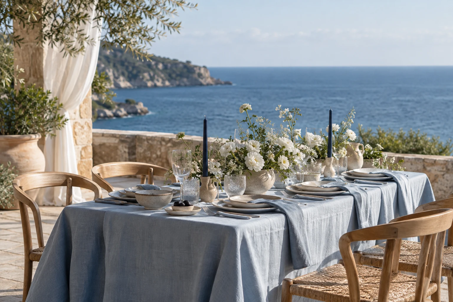 Editorial concept of a seaside terrace table with blue linen, white flowers, woven chairs and an ocean view